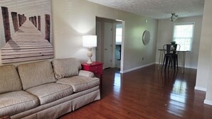 A living room with a wooden floor, a beige sofa, a red nightstand, a lamp, a wall-mounted mirror, and a dining area with a table and chairs.