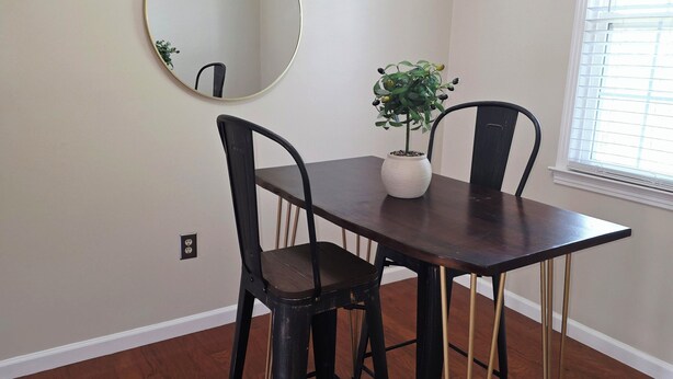 A dining area with a table, chairs, a potted plant, a round mirror, and a window with blinds.
