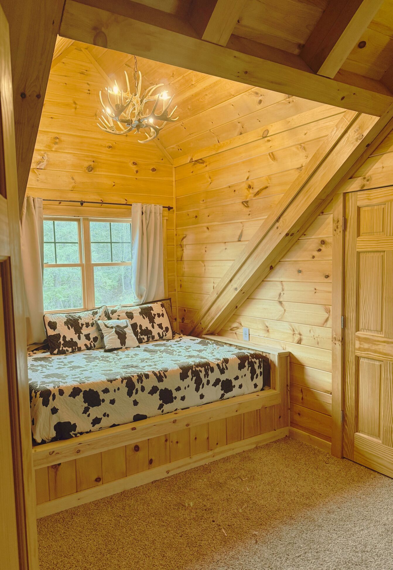 A cozy cabin bedroom with a sloped ceiling, a bed with a cow print bedspread, a window with curtains, and a wooden door.