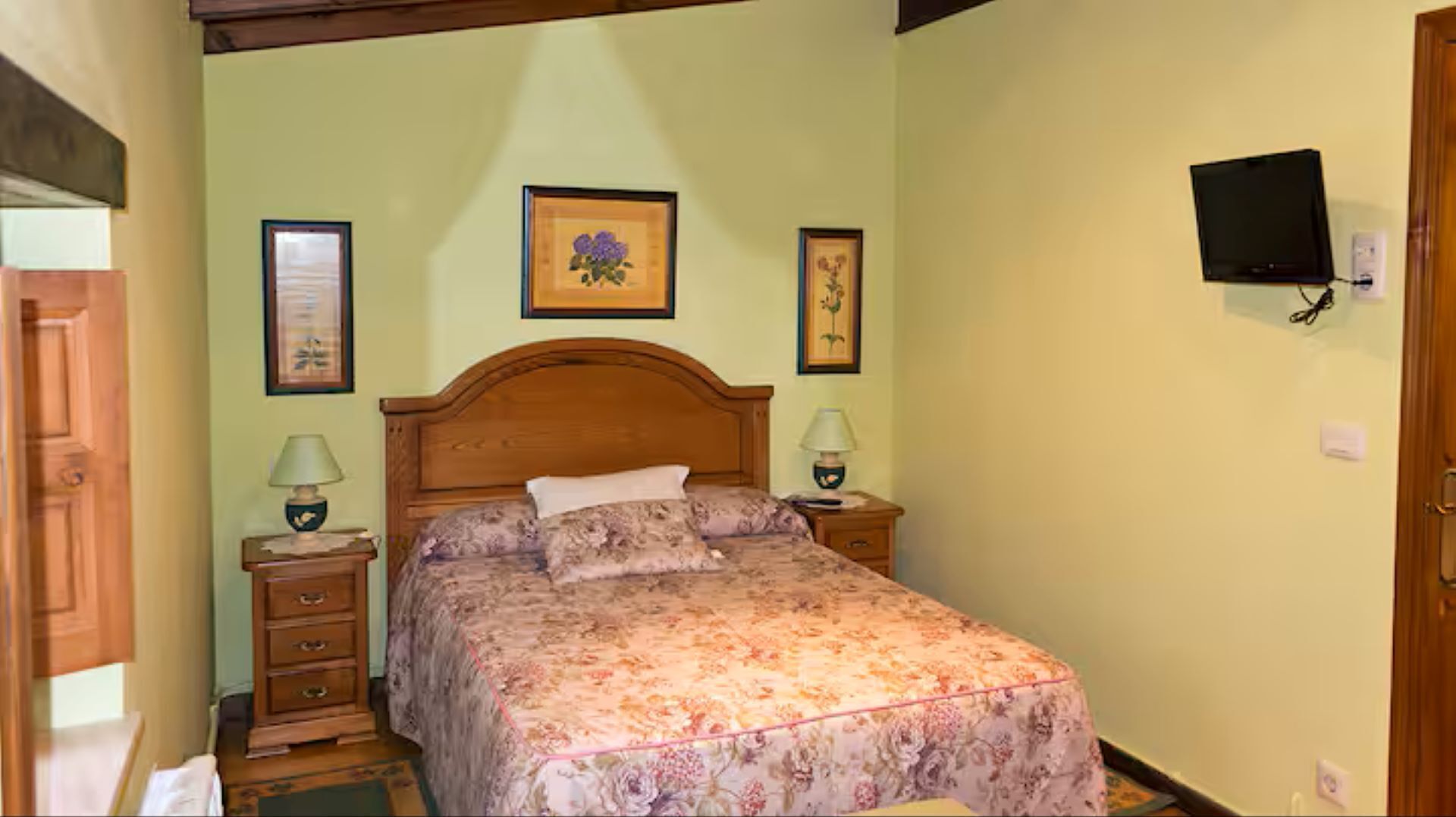 A bedroom with a wooden bed, two bedside tables, a television mounted on the wall, and framed artwork on the walls.