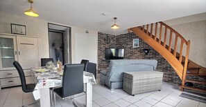 Un área de comedor con una mesa preparada para cuatro personas, un sofá gris, un reposapiés de mimbre, una escalera de madera y una pared de ladrillo decorativa.