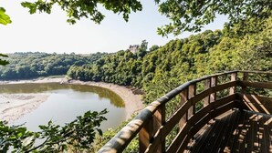 Een houten terras met uitzicht op een rivier, een bebost glooiend landschap en een kasteelachtige structuur in de verte.