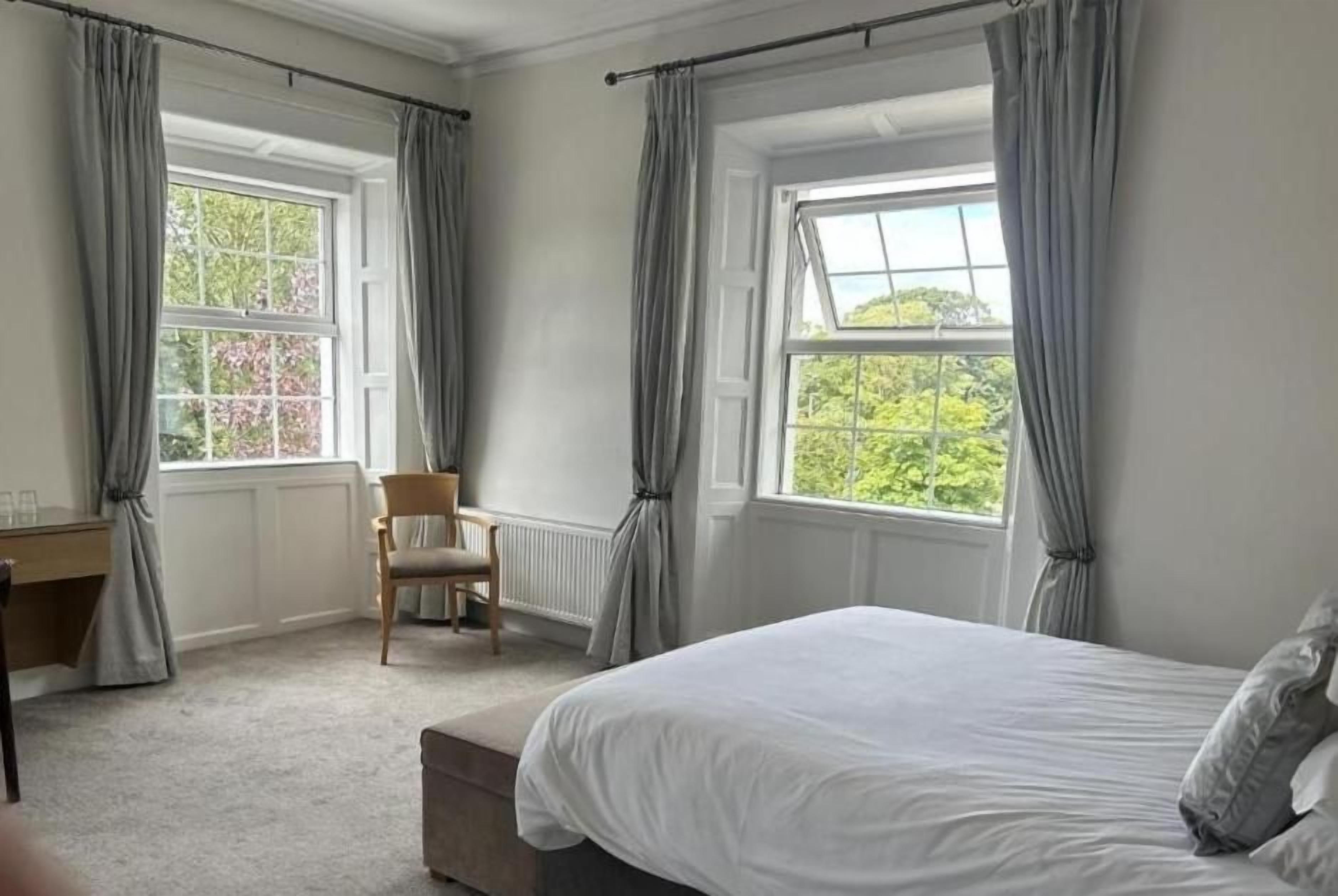 Un dormitorio con una cama, una silla, un escritorio y dos ventanas con cortinas.