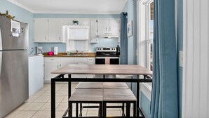 A compact kitchen with white cabinets, a stainless steel refrigerator, a wooden table, and a window with blue curtains. - UNIT C: Walk to Beach/Intercoastal/Fishing Pier (Holly Ridge)