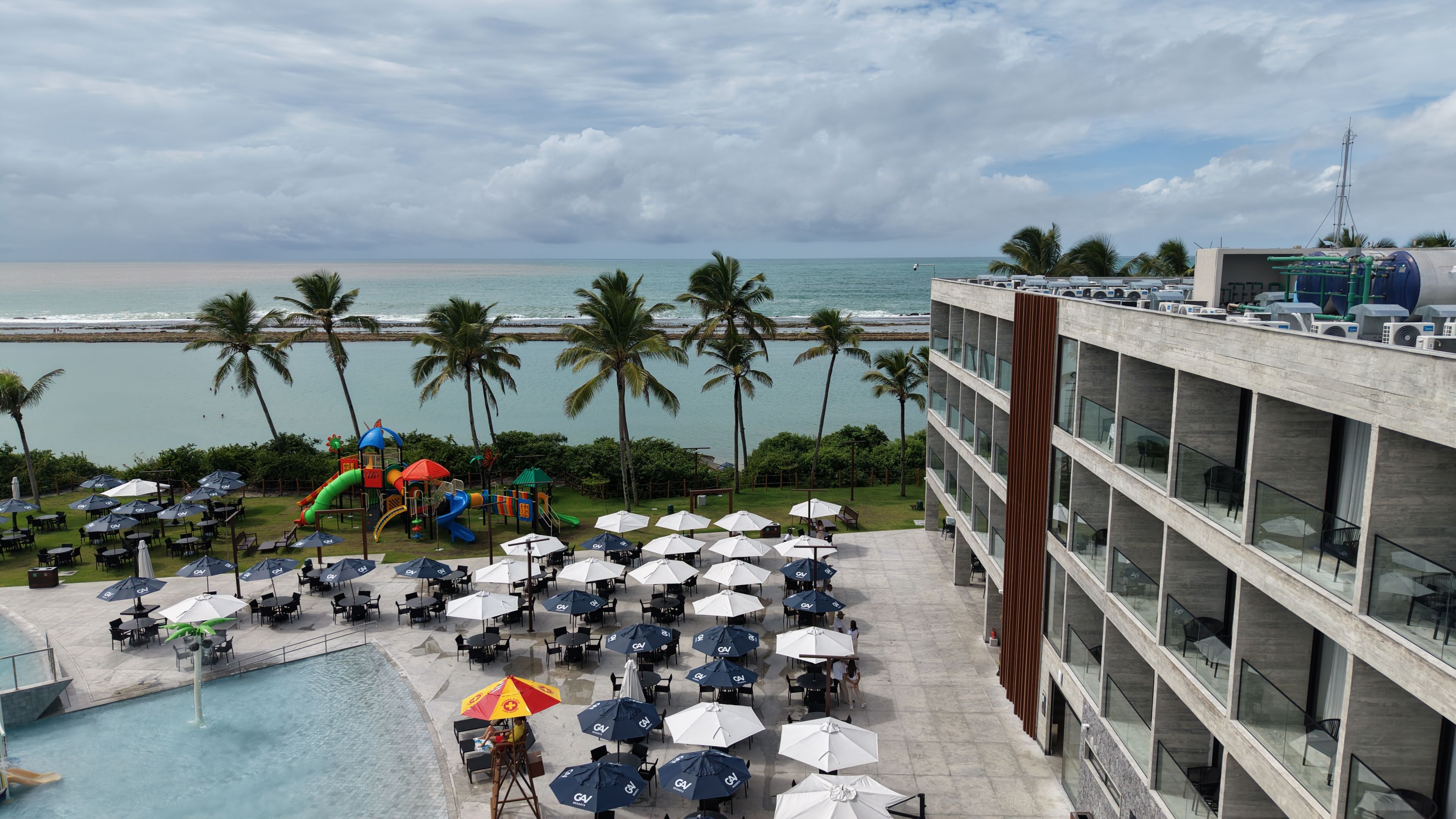 Ein Hotelpoolbereich mit Sonnenschirmen, Liegestühlen und einer Spielanlage für Kinder.