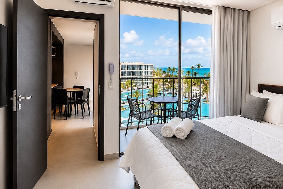 A hotel room with a bed, a balcony with a view of the pool and ocean, and a dining area.