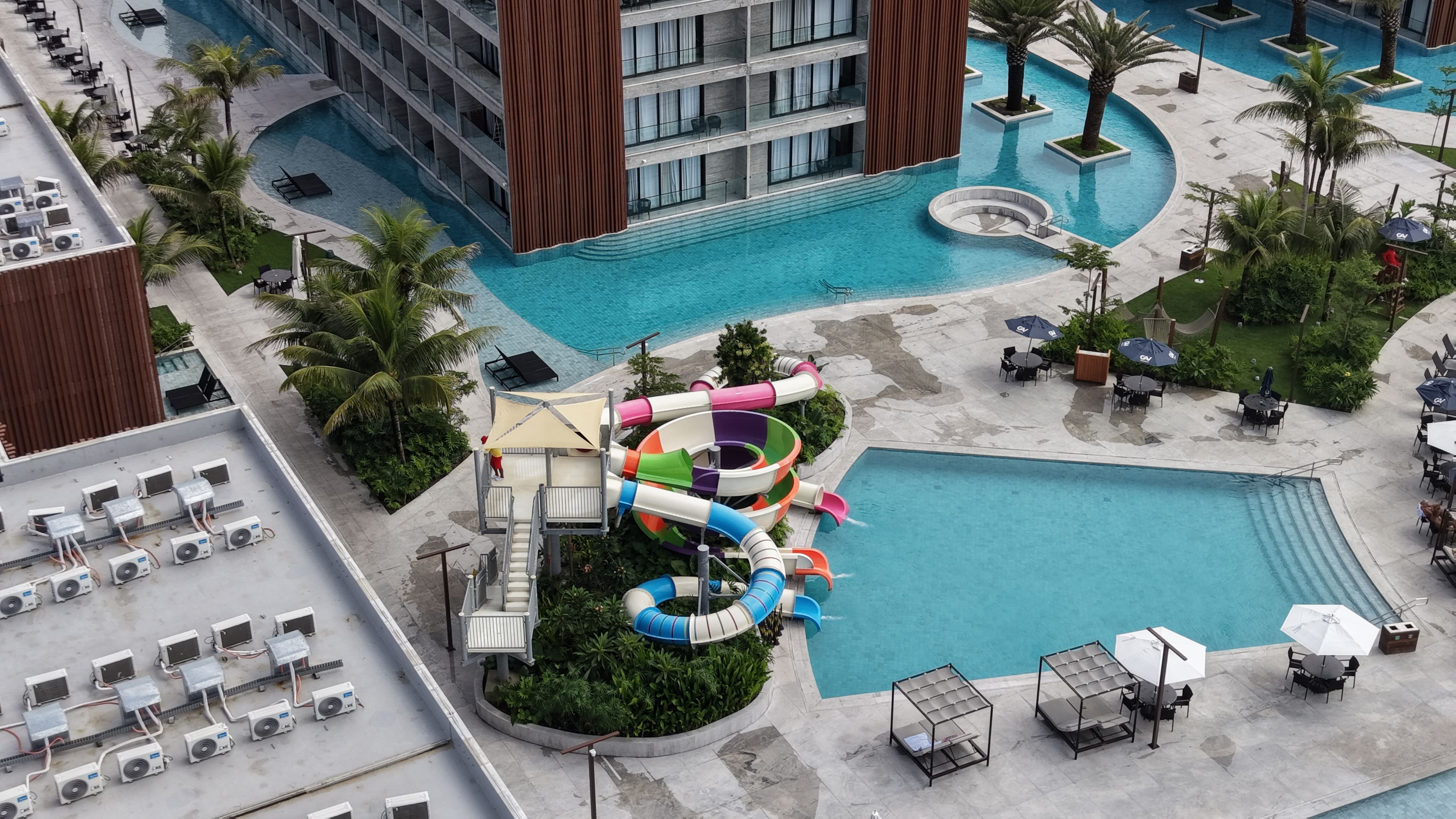 Zona de piscina de hotel con tobogán de agua, sillas de descanso y un edificio con balcones.