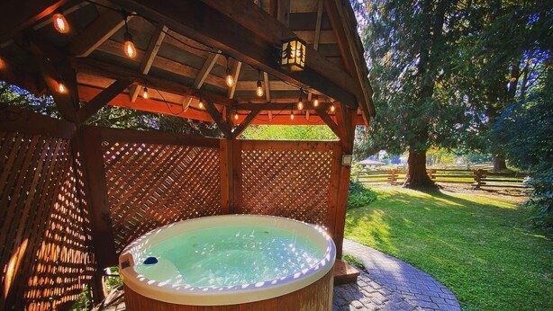 A hot tub under a wooden pergola with a lattice wall, surrounded by a brick patio and a grassy area. - "Darla" the Glamping Trailer at Ashford Lodge (Ashford)