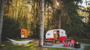 A forest campsite with a small cabin, a camper, picnic tables, chairs, and a fire pit. - "Darla" the Glamping Trailer at Ashford Lodge (Ashford)