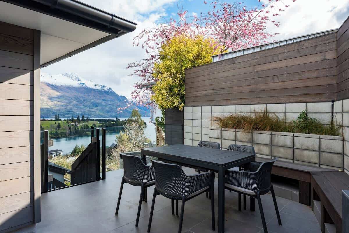 Een moderne eethoek buiten met een tafel en stoelen, uitzicht op een rivier en bergen, en een houten muur begroeid met groen.