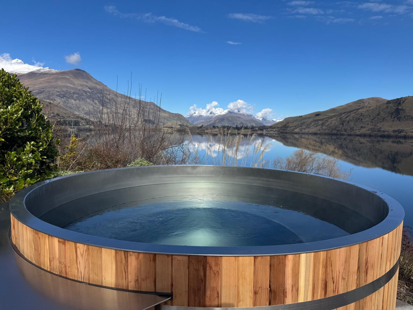 A hot tub with a wooden exterior, surrounded by a natural landscape with mountains and a lake.
