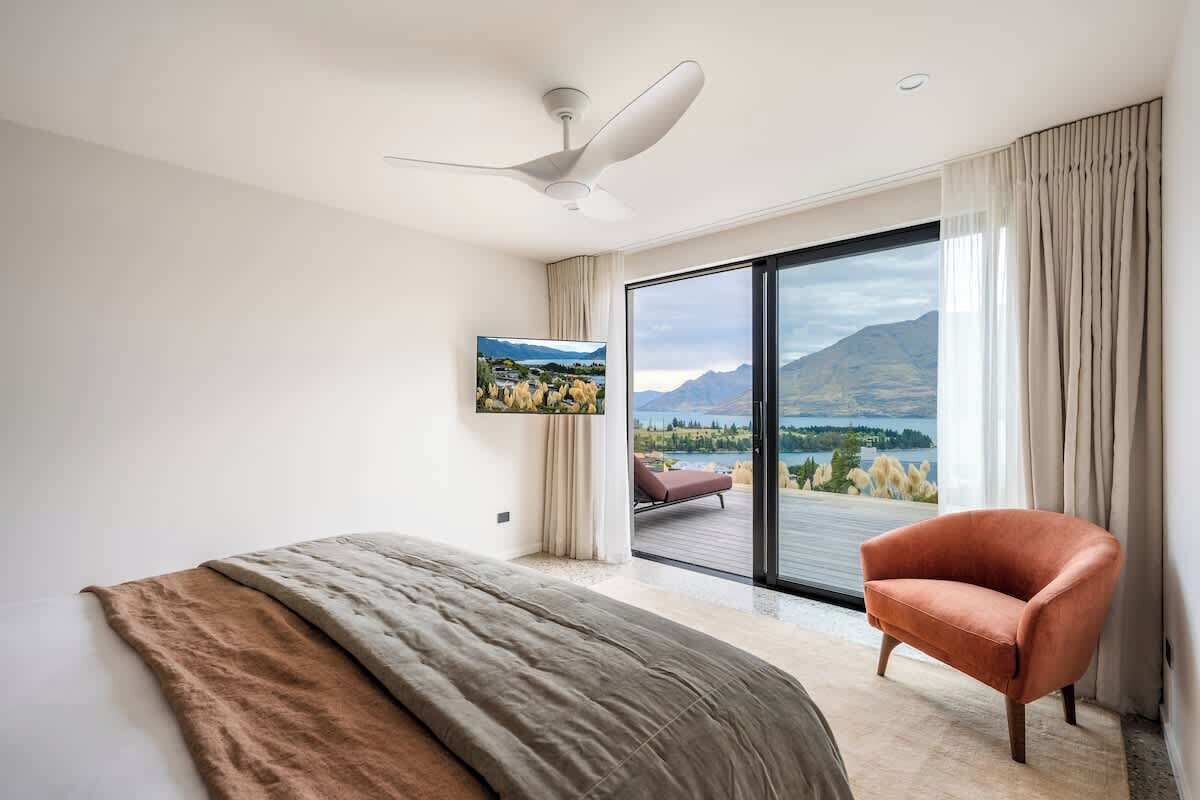 A modern hotel room with a bed, a ceiling fan, a large window with a view, and a single armchair.