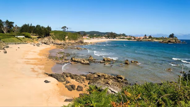 Une plage côtière aux eaux cristallines, avec un rivage rocheux et des bâtiments visibles au loin.