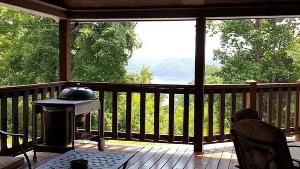 En overdækket terrasse udendørs med grill, et bord med stole og udsigt til sø og bjerge.