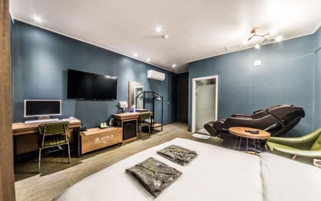 A hotel room with a bed, a desk with a computer, a TV, a massage chair, and a small table.