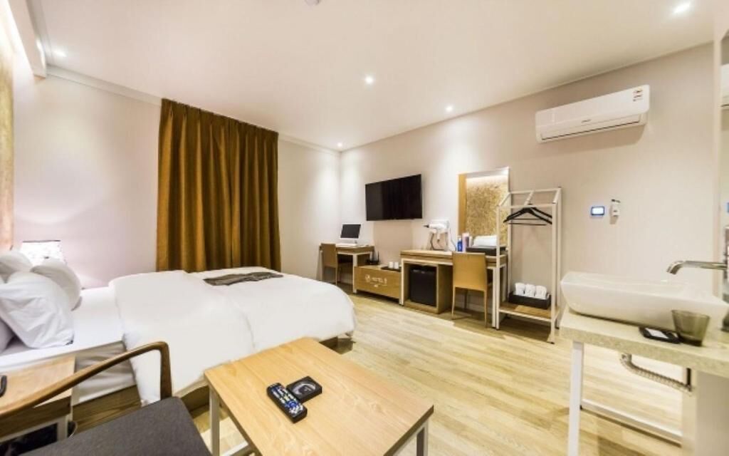 A hotel room with a bed, a desk, a chair, a table, a TV, a wardrobe, a bathroom with a sink, and an air conditioning unit.