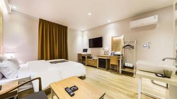 A hotel room with a bed, a desk, a chair, a table, a TV, a wardrobe, a bathroom with a sink, and an air conditioning unit.