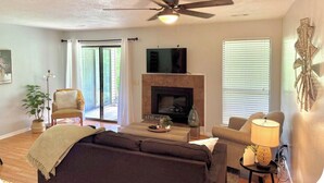 Una sala de estar con chimenea, ventilador de techo, televisor y un ventanal con persianas.