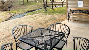 Un patio con una mesa y sillas de metal, un banco y vistas a un arroyo y árboles.