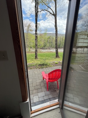 A view through a glass door to a patio with a red chair and a grassy area with trees.