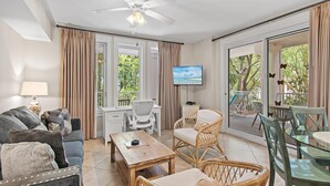 Ein Wohnzimmer mit einer Couch, einem Couchtisch, Korbstühlen, einem Fernseher und einem Balkon mit Gartenmöbeln.