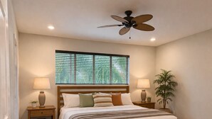 Une chambre à coucher comprenant un lit, un ventilateur de plafond, deux tables de chevet avec des lampes, une fenêtre avec des stores et une plante en pot.