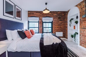 A modern bedroom with a brick wall, a bed with white bedding, red and black pillows, a hanging light fixture, and a view of buildings through large windows. - Modern 2-bed Flat in Central Leeds - Sleeps 6 (Leeds)
