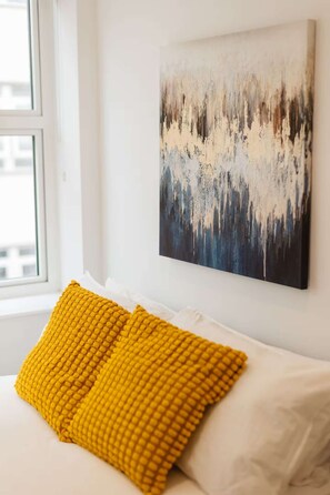 A bed with white bedding and a single yellow pillow, a painting on the wall, and a window with a view of buildings. - Central Leeds 1-bed Apartment Near Train Station (Leeds)
