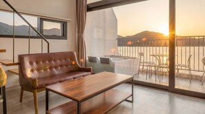 Ein modernes Zimmer mit Ledersofa, Holztisch und einem Balkon mit Blick auf Sonnenuntergang und Berge.