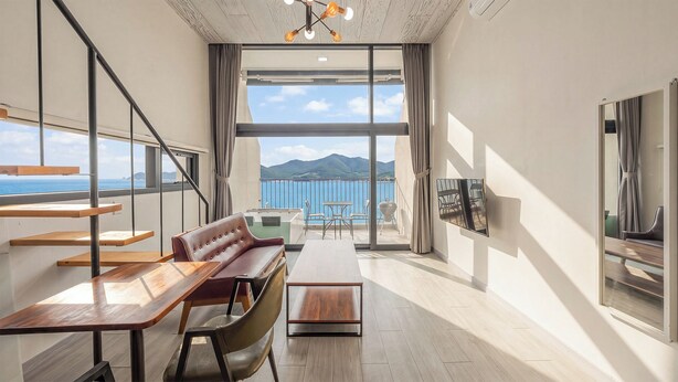 Un intérieur moderne avec un canapé, une table à manger et des chaises, de grandes fenêtres offrant une vue sur la mer et les montagnes, et un luminaire au plafond.