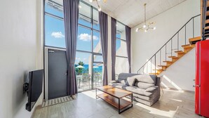 Un salon moderne avec un canapé, une table basse et un téléviseur. On y trouve un escalier avec un mur d’accent rouge et une vue sur la mer grâce à de grandes fenêtres.