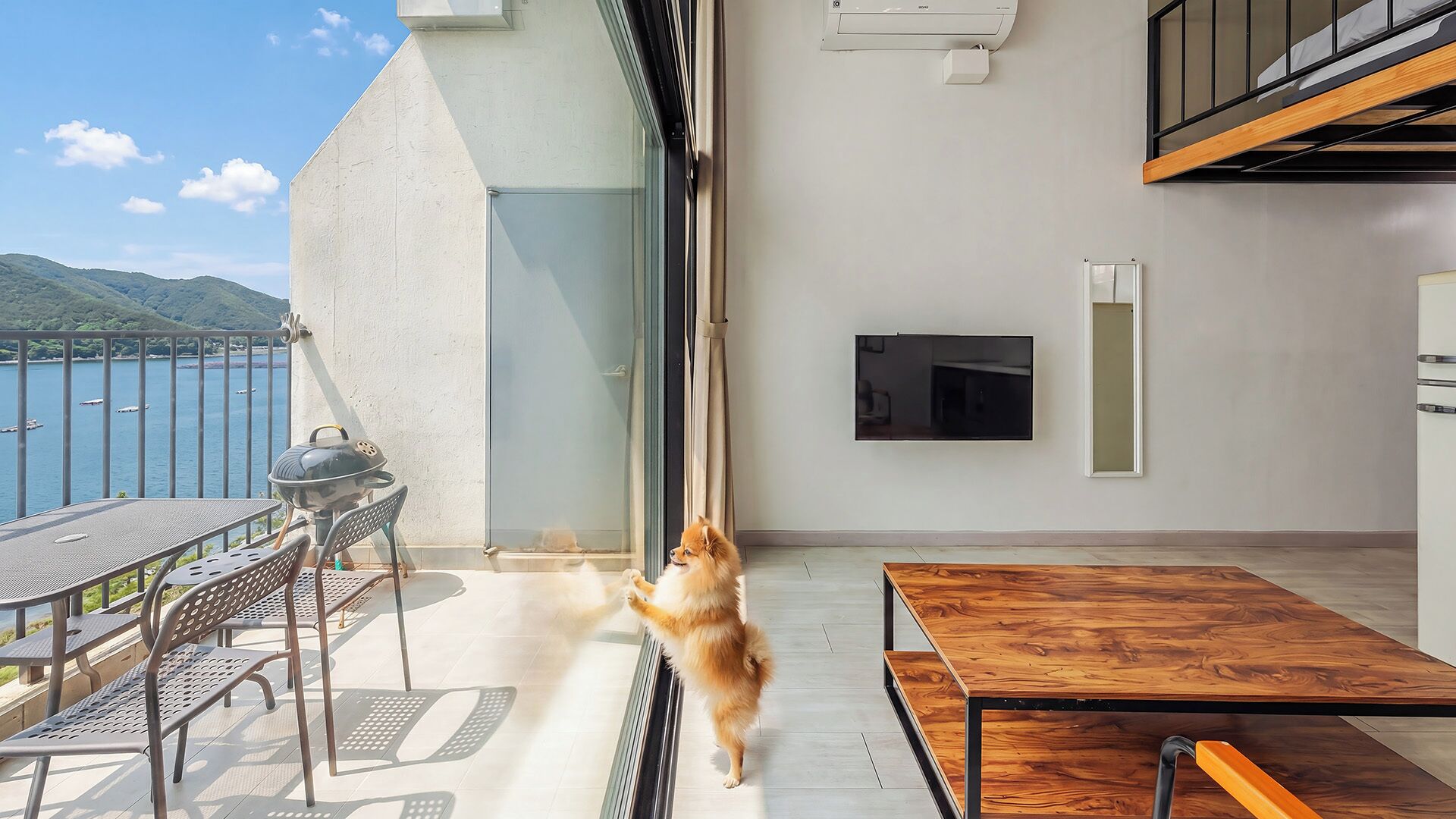 Een balkon met een tafel, stoelen en een hond die bij een schuifdeur staat.