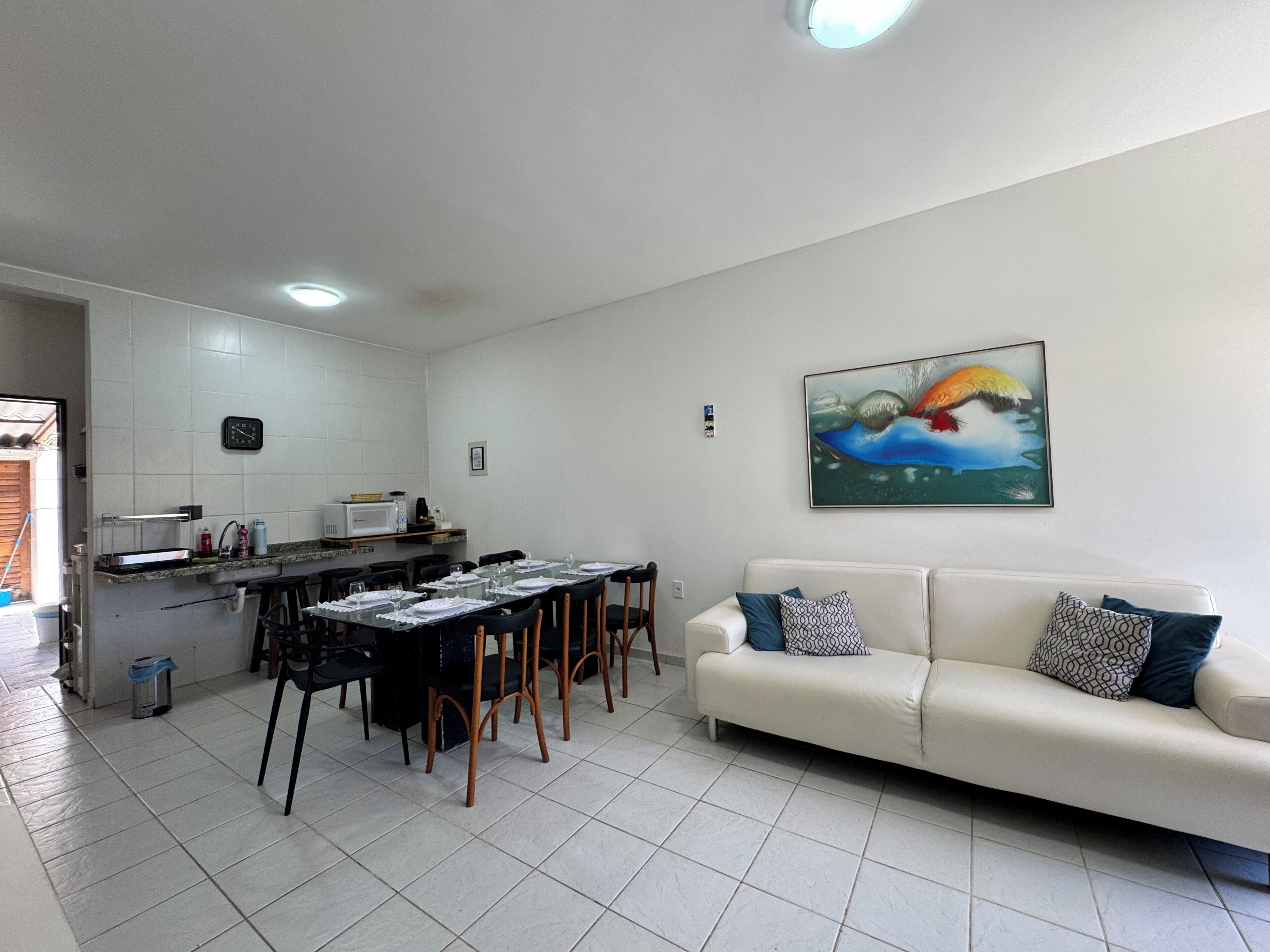 A modern living room with a dining area, white walls, a sofa with patterned cushions, a painting, and a kitchen area with a microwave.