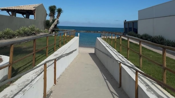 Ein Weg führt zu einem Strand mit blauem Himmel, Palmen und einem Gebäude mit einem Ziegeldach.