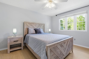 Un dormitorio con cama, mesitas de noche, ventilador de techo y ventana con vista a árboles.