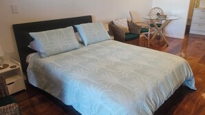 Un lit bien fait, recouvert d’une couvre-lit à motifs, une chaise en osier avec un coussin, une petite table ronde avec un bol, et un coin cuisine avec des placards et un évier.