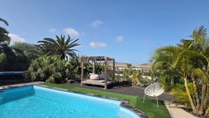 Uma piscina com deck de madeira, um gazebo com telhado de palha e vegetação tropical exuberante.