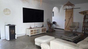 Uma sala de estar com uma TV de tela plana, um sofá branco, uma mesa de centro de madeira, um armário branco e uma cadeira rústica.
