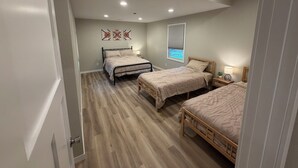 A bedroom with two beds, a window with blinds, a bedside table with a lamp, and a wall-mounted artwork.