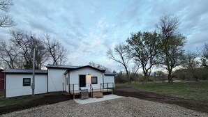 A single-story mobile home with a front porch, a small attached garage, and a gravel driveway.
