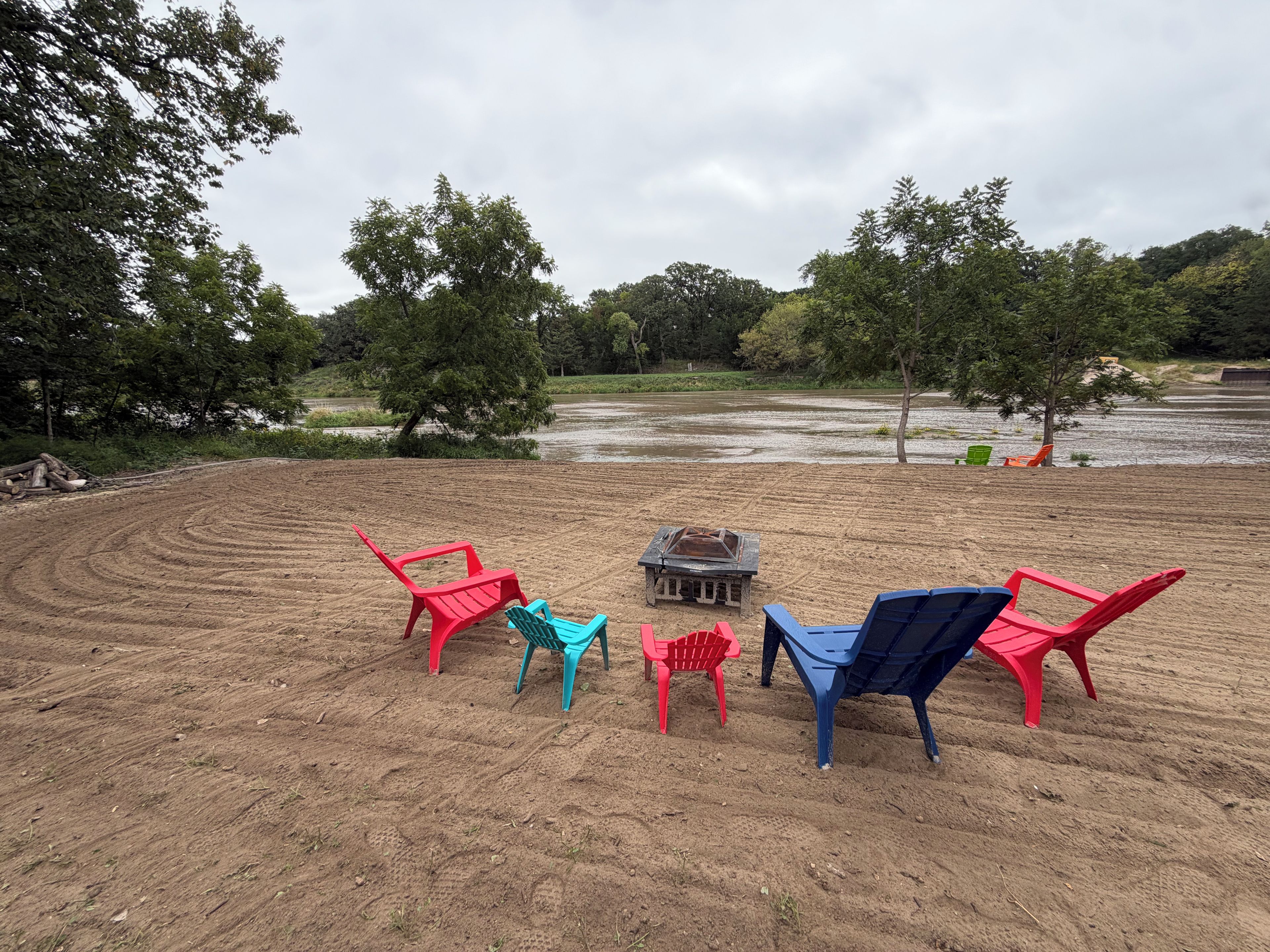 Un espace sablonneux avec plusieurs chaises en plastique de couleurs vives et une petite table, entouré d’arbres et bordé par une étendue d’eau.