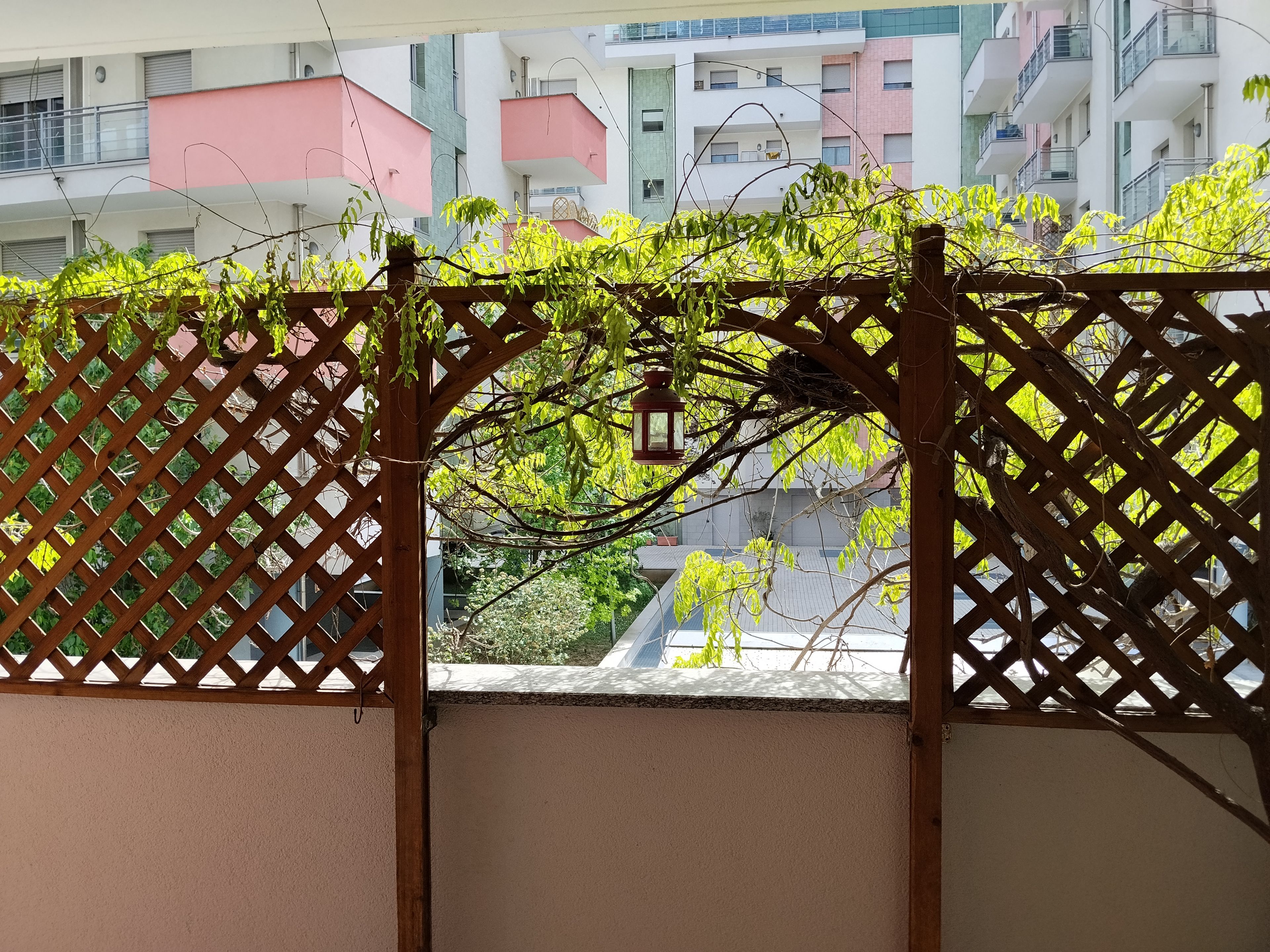 Une porte en treillis de bois ornée d’une lanterne, qui donne sur un balcon offrant une vue sur des immeubles à plusieurs étages.