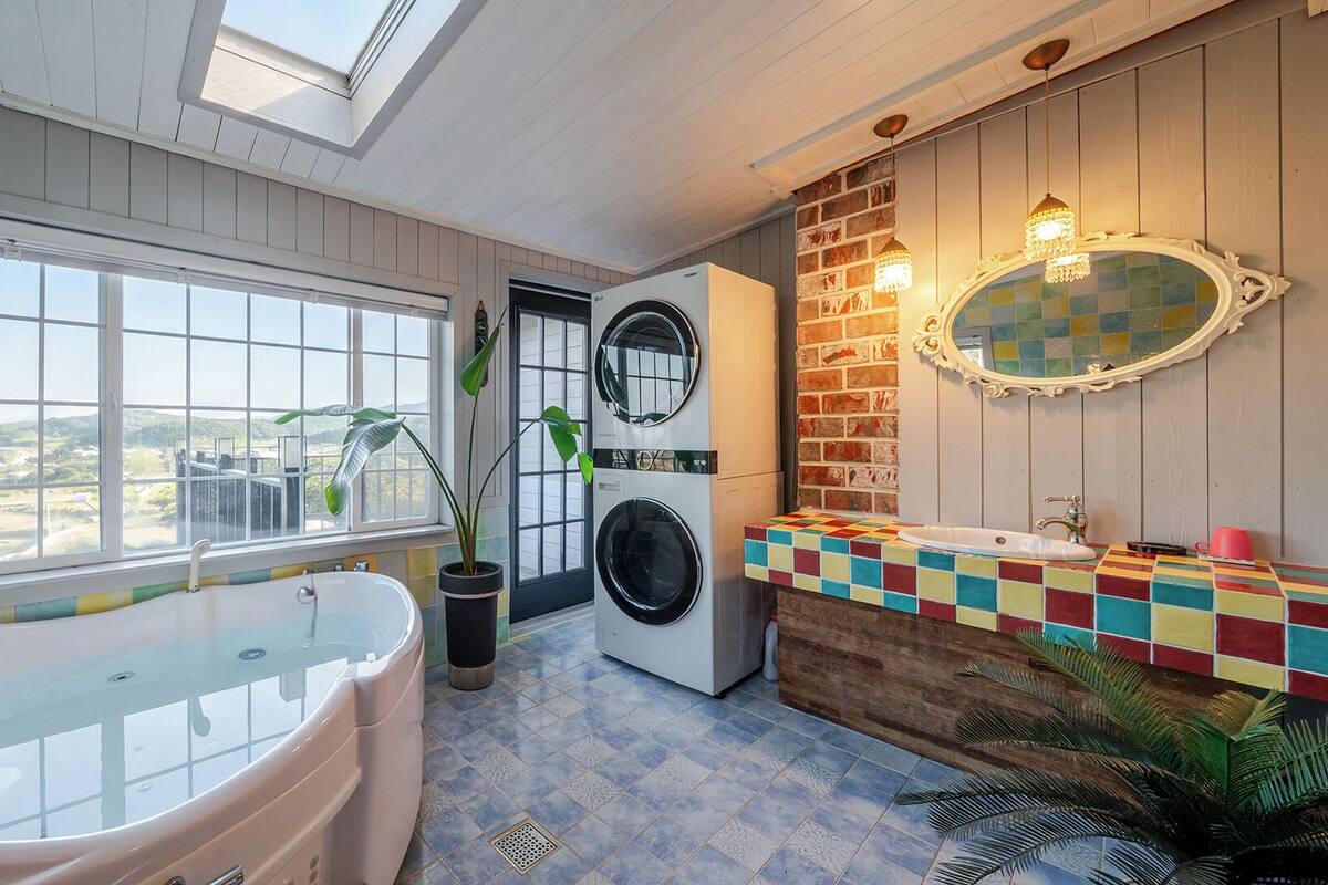 Un baño con bañera, lavadora, lavamanos y un revestimiento de azulejos de colores, además de un ventanal grande.