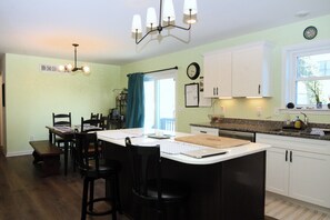 A kitchen with a central island, white cabinets, a dining area with chairs, a chandelier, and a window with curtains. - Rehoboth Beach House Pet Friendly Fenced Yard Next to Trail (Rehoboth Beach)