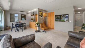 A modern living room with a sofa, a dining area with a table and chairs, a kitchen with wooden cabinets, and a large window. - Roberts Street Retreat - Taupo CBD (Taupo)