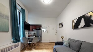 A living room with a grey sofa, a dining table with chairs, a kitchen area, and a white door.
