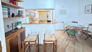 A compact kitchen with a central island, two bar stools, a dining table with chairs, and a bookshelf with books and decorative items. - Domus Floris Positano (Positano)