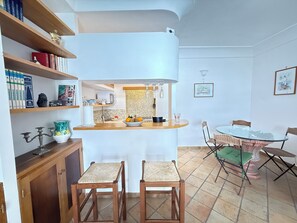 Una cocina compacta con una isla central, dos taburetes de bar, una mesa de comedor con sillas y una estantería con libros y adornos.