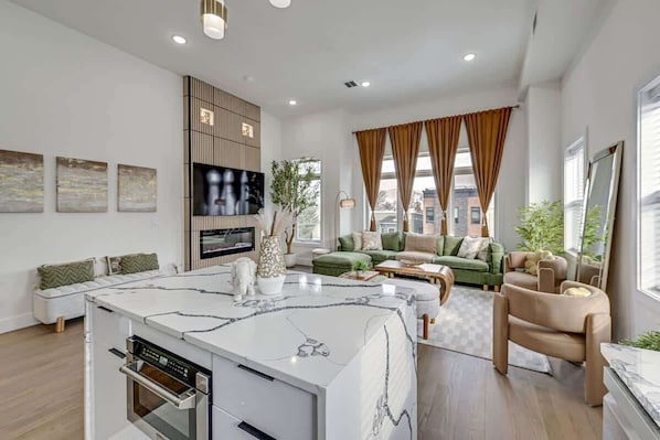 A modern living room with a kitchen island, a flat-screen TV, and a green sofa set. - Luxury Mansion, Rooftop, Parking, Near NYC & EWR (Jersey City)