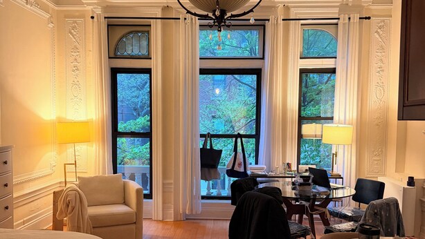 Ein Hotelzimmer mit Bett, einer Sitzecke mit Tisch und Stühlen, einer Kristallleuchter, großen Fenstern mit Vorhängen und Blick auf Bäume.
